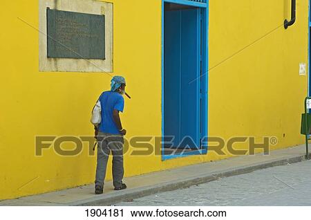 Stock Image - Man Smoking Cigar, Walking Along Yellow Building, Rear View, Havana, Cuba. Fotosearch