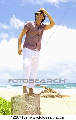 Stock Image - Man standing on a tree stump on the beach; Kealia, Kauai, Hawaii, United States of America. Fotosearch