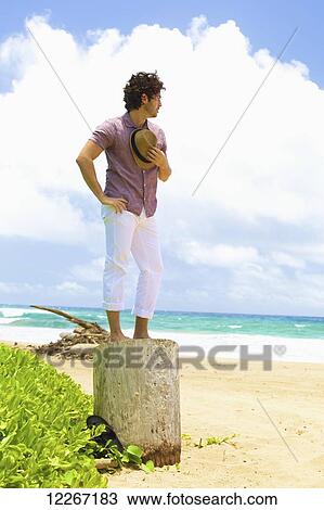 Stock Image - Man standing on a tree stump on the beach; Kealia, Kauai, Hawaii, United States of America. Fotosearch