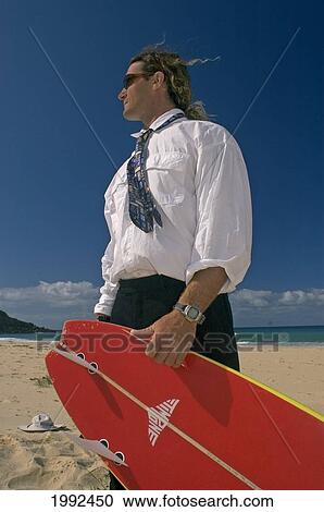 Stock Image - Man Standing On Beach In Business Suit Holding A Surfboard.. Fotosearch