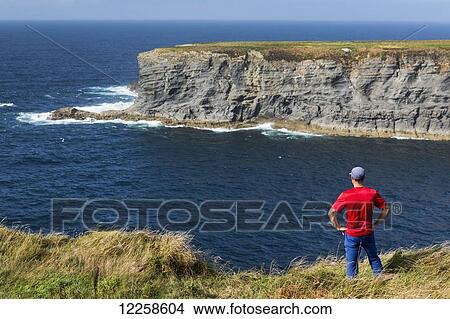 Picture - Man standing on grassy cliff's edge overlooking large rock island in the ocean with blue sky; Kilkee, County Clare, Ireland. Fotosearch
