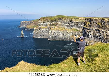 Man standing on grassy cliff's edge overlooking large sea stack rock structure in the ocean with boat taking a photo with phone; Liscannor, County Clare, Ireland View Large Photo Image Stock Image - Man standing on grassy cliff's edge overlooking large sea stack rock structure in the ocean with boat taking a photo with phone; Liscannor, County Clare, Ireland. Fotosearch