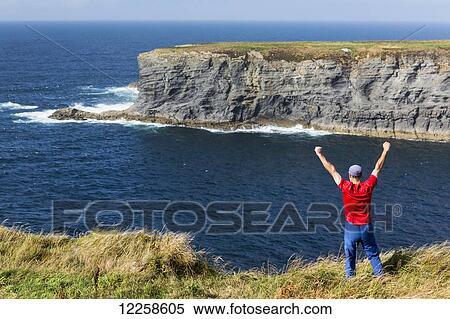Man standing on grassy cliff's edge with arms raised overlooking large rock island in the ocean with blue sky; Kilkee, County Clare, Ireland View Large Photo Image Stock Photography - Man standing on grassy cliff's edge with arms raised overlooking large rock island in the ocean with blue sky; Kilkee, County Clare, Ireland. Fotosearch