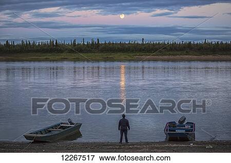 Stock Photo - Man standing on the banks of the Porcupine River with full moon reflection; Old Crow, Yukon. Fotosearch