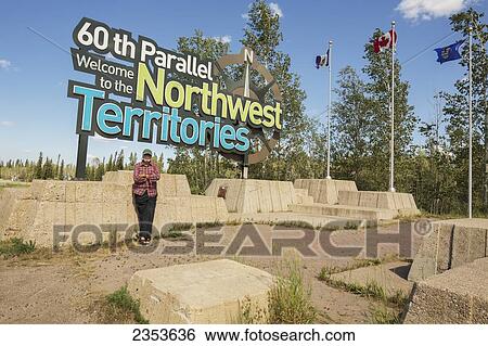 Stock Photograph - Man Standing Under The Northwest Territories/Alberta Border Sign; Northwest Territories, Canada. Fotosearch