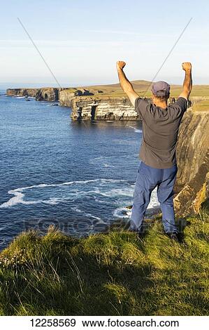 Man standing with arms raised in the air on grassy cliff edge overlooking the dramatic rock cliffs along the shoreline at sunset; Kilkee, County Clare, Ireland View Large Photo Image Stock Photo - Man standing with arms raised in the air on grassy cliff edge overlooking the dramatic rock cliffs along the shoreline at sunset; Kilkee, County Clare, Ireland. Fotosearch
