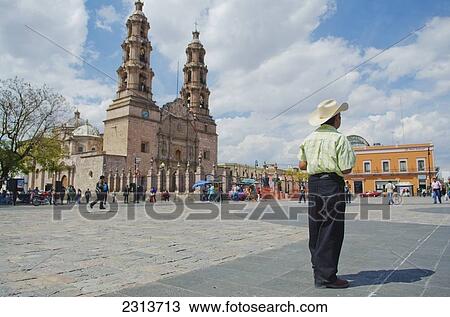 Stock Image - Man wearing cowboy hat standing in large city plaza beside large cathedral; Aguascalientes, Aguascalientes state, Mexico. Fotosearch