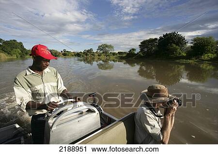 Stock Image - Man with camera and tour guide driving boat down a tranquil river in Lower Zambezi National Park; Zambia. Fotosearch