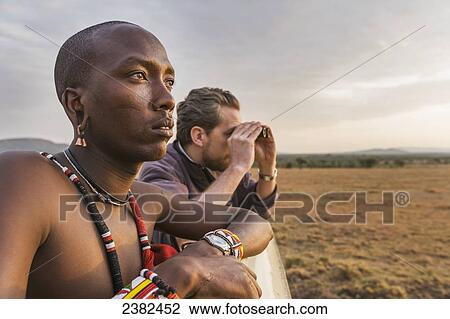 Stock Image - Masai tribesman and a tourist look for wildlife in the early morning in the Masai Mara; Kenya. Fotosearch