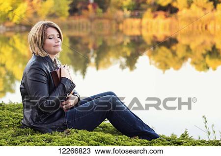 Stock Image - Mature Christian woman embracing her Bible in a city park beside a lake in autumn; Edmonton, Alberta, Canada. Fotosearch