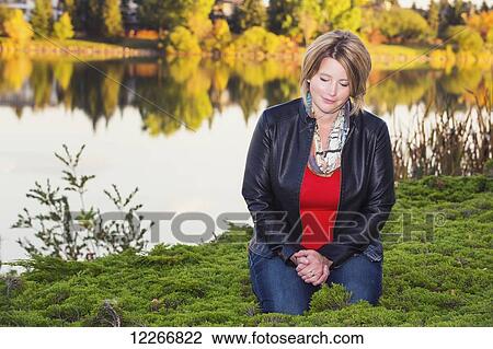 Stock Image - Mature Christian woman kneeling and praying beside a lake in autumn; Edmonton, Alberta, Canada. Fotosearch