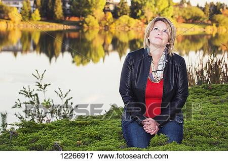 Stock Photo - Mature christian woman kneeling and praying beside lake in autumn; Edmonton, Alberta, Canada. Fotosearch