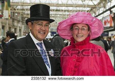 Stock Image - Mature Couple Waiting At Waterloo Station For Train To Royal Ascot On Lady's Day; London, England, Uk. Fotosearch