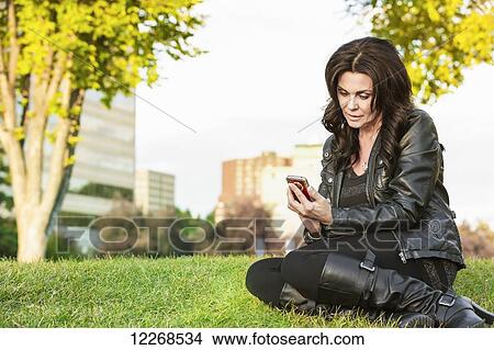 Picture - Mature woman texting on her smart phone in an outdoor urban park in autumn; Edmonton, Alberta, Canada. Fotosearch