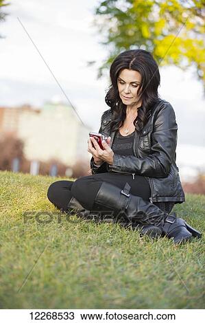 Stock Image - Mature woman texting on her smart phone in an outdoor urban park in autumn; Edmonton, Alberta, Canada. Fotosearch