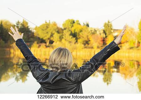 Stock Image - Mature woman with arms raised in worship beside lake in autumn; Edmonton, Alberta, Canada. Fotosearch