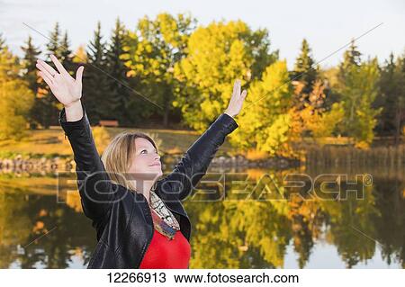 Stock Image - Mature woman with arms raised in worship beside lake in autumn; Edmonton, Alberta, Canada. Fotosearch