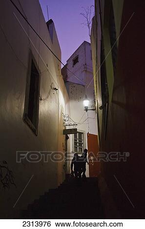 Stock Photograph - Mexico, Guanajuato, Guanajuato, Silhouettes of people walking up narrow alley at night. Fotosearch