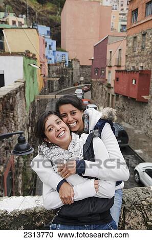 Stock Image - Mexico, Guanajuato State, Guanajuato, Two young women hugging and laughing in downtown area. Fotosearch