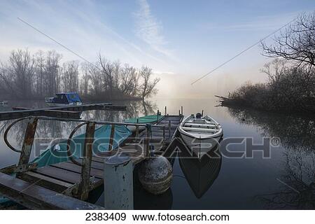Stock Photo - Mist rising over Lake Maggiore with a canoe tethered to a dock; Locarno, Ticino, Switzerland. Fotosearch