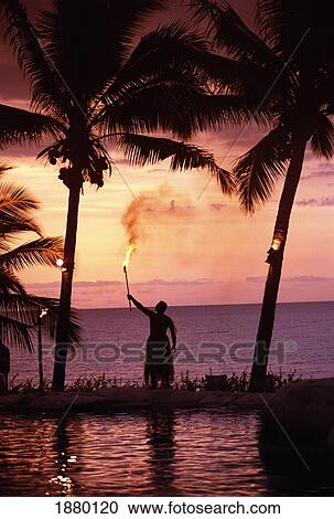 Stock Image - Native In A Grass Skirt Holding A Flaming Torch By Coast At Sunset. Fotosearch