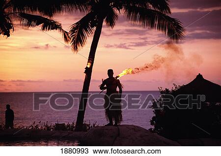 Stock Image - Native In A Grass Skirt Holding A Flaming Torch By Coast At Sunset. Fotosearch