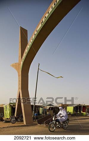 Stock Image - Niger, Sahara Desert, Agadez Region, Veiled Tuareg riding bike crossing city main gate; Agadez. Fotosearch