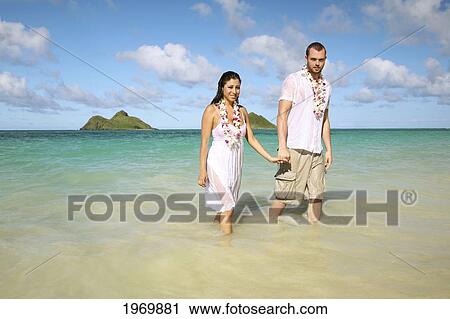 Stock Image - Oahu, Hawaii, Lanikai, Young Couple Both Holding Hands And Walking Out Of Water.. Fotosearch