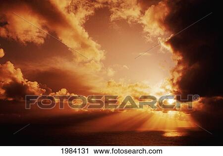Stock Image - Orange Sunset Over Ocean, Sunrays Bursting Through Puffy Clouds.. Fotosearch