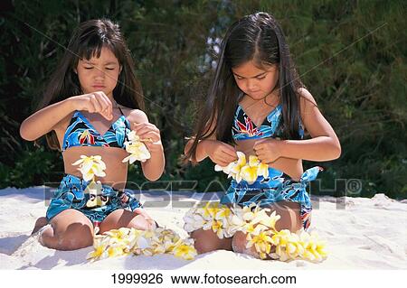 Stock Photograph - Pai Of Hawaiian Girls Wear Matching Outfits On Beach, Stringing Plumeria Flowers C1485. Fotosearch