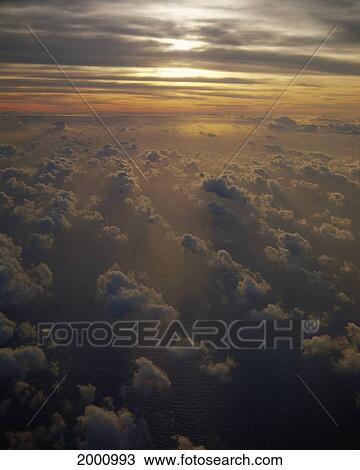 Stock Image - Pale Orange Sunset From Plane Large Puffy Clouds, Sun Streaks Through Sky. Fotosearch