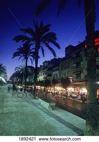 Stock Image - Palm Tree Lined Promenade With People Eating In Restaurants At Dusk. Fotosearch
