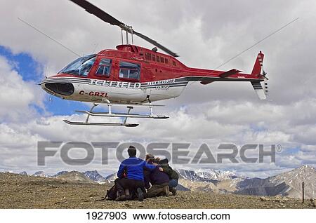 Stock Image - Passengers Watching Helicopter Fly Off Crouching On Mountain Ridge, Alberta, Canada. Fotosearch