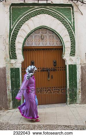 Stock Image - Pedestrian dressed in brightly colored clothing walking by ornate doorway in Quartier Habbous; Casablanca, Morocco. Fotosearch