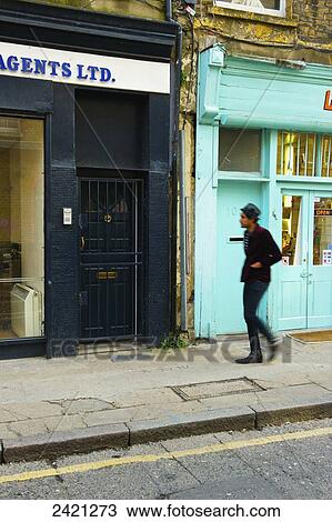 Stock Image - Pedestrian walking along street beside retail buildings, Brick Lane; London, England. Fotosearch