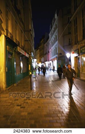Stock Image - Pedestrians on a walkway outside retail buildings at nighttime; Paris, France. Fotosearch