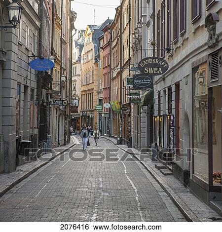 Stock Photograph - Pedestrians Walking Down A Street With Shop Signs Mounted On The Sides Of Buildings; Stockholm Sweden. Fotosearch
