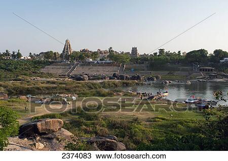 Stock Image - People and boats along the shore of the river with buildings in the background; Hampi, Karnataka, India. Fotosearch