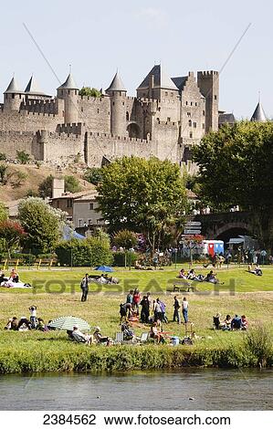 Stock Image - People gathering on the lawn by the river with castle and ramparts in the background; Carcassonne, Languedoc-Rousillion, France. Fotosearch