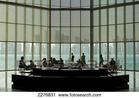 Stock Image - People sitting at tables in front of large window in the Museum of Islamic Art with view of the modern city skyline of Doha; Doha, Qatar. Fotosearch