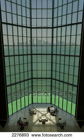 Stock Image - People sitting at tables in large window of the Museum of Islamic Art with view of modern skyline of Doha behind, Doha, Qatar.. Fotosearch
