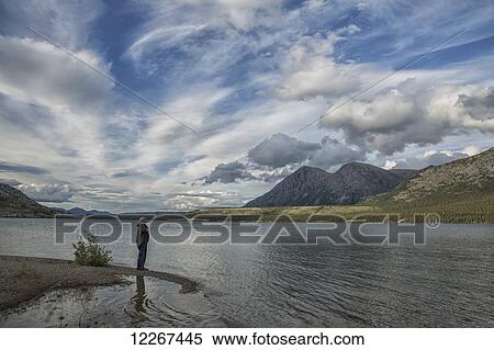 Stock Photography - Person standing on the shores of Kusawa Lake as the day is slowly coming to an end; Yukon, Canada. Fotosearch