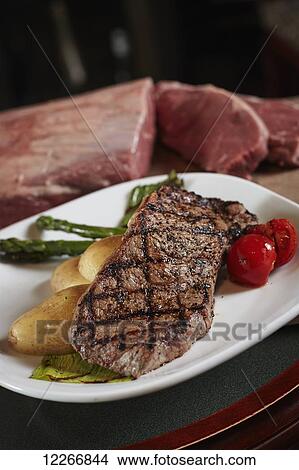 Picture - Plated dinner of New York striploin with roasted tomato, potato and asparagus in front of raw cut of beef. Fotosearch