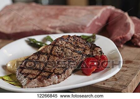 Stock Photography - Plated dinner of New York striploin with roasted tomato, potato and asparagus in front of raw cut of beef. Fotosearch