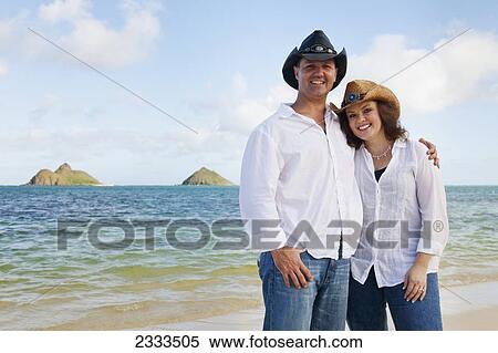 Stock Photography - Portrait of a couple in white shirts and western hats standing on the beach; Honolulu, Oahu, Hawaii, United States of America. Fotosearch