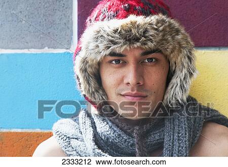 Stock Image - Portrait of a shirtless young man wearing a scarf and fur trimmed hat standing against a colourful wall; Honolulu, Oahu, Hawaii, United States of America. Fotosearch