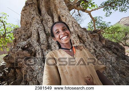 Stock Photo - Portrait of a young girl standing in front of a large tree; Ethiopia. Fotosearch