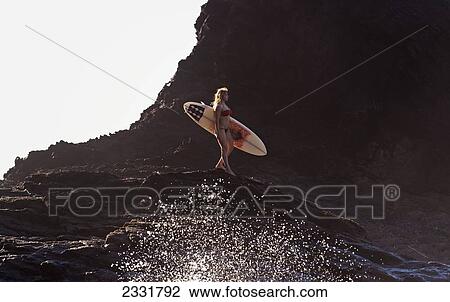 Stock Image - Portugal, Algarve, Carrapateira, Distant View Of Female Surfer With Surfboard Standing On Rock; Playa Do Amado. Fotosearch