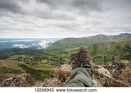 Stock Image - POV of hikers legs and feet as he rests and enjoys the view from Flat Top Mountain overlooking the Anchorage bowl, Southcentral Alaska, Summer. Fotosearch