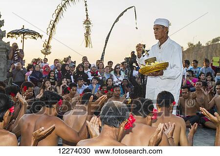 Stock Image - Priest carrying incense and water for blessings surrounded by men sitting in a circle chanting in trance during a Kecak dance performance, Ulu Watu, Bali, Indonesia. Fotosearch